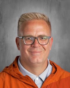 Smiling man with glasses, short blond hair, wearing an orange jacket and striped shirt, against a gray background.