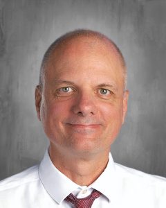 Bald man smiling, wearing a white shirt and red tie, with a gray background.