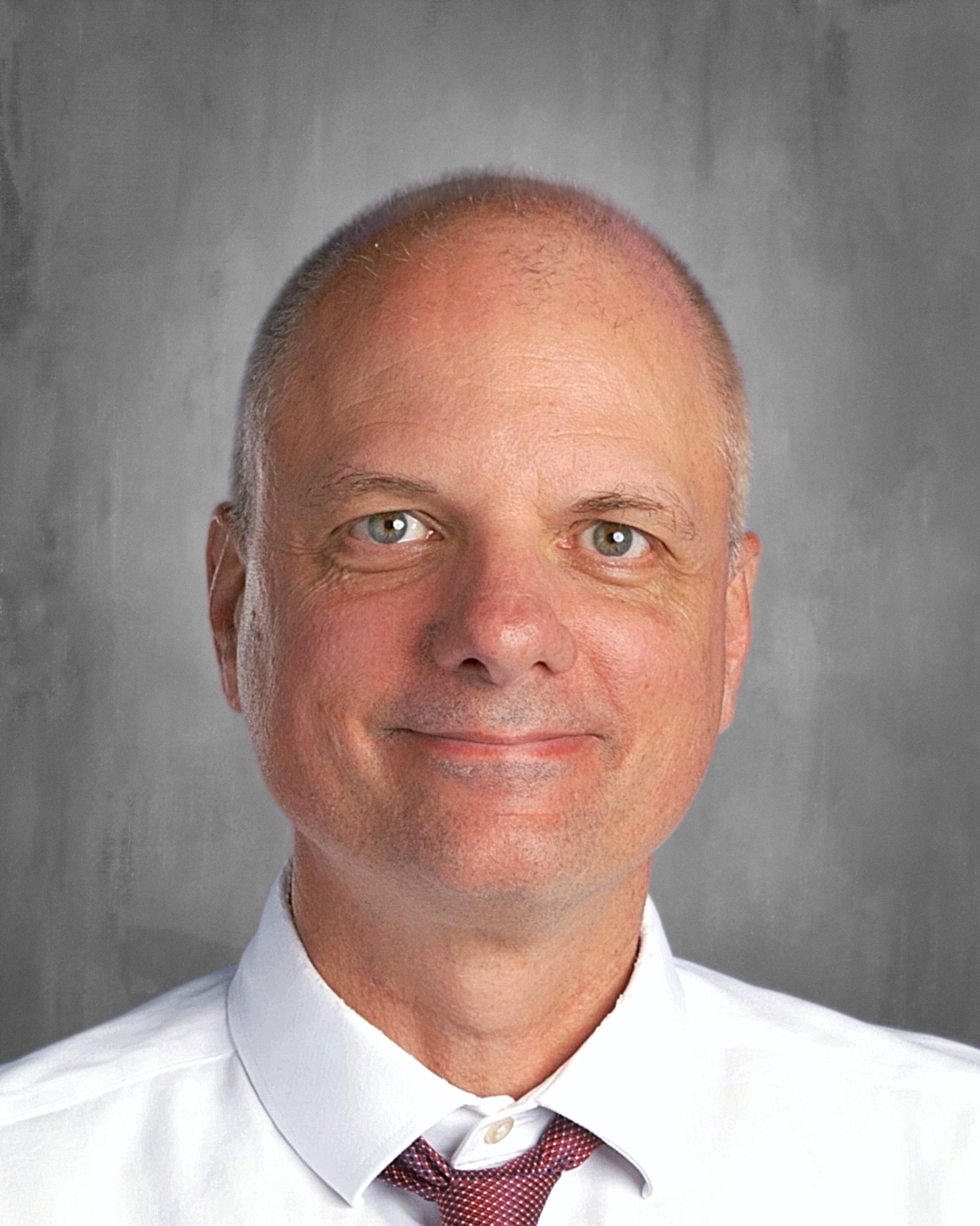 Bald man smiling, wearing a white shirt and red tie, with a gray background.