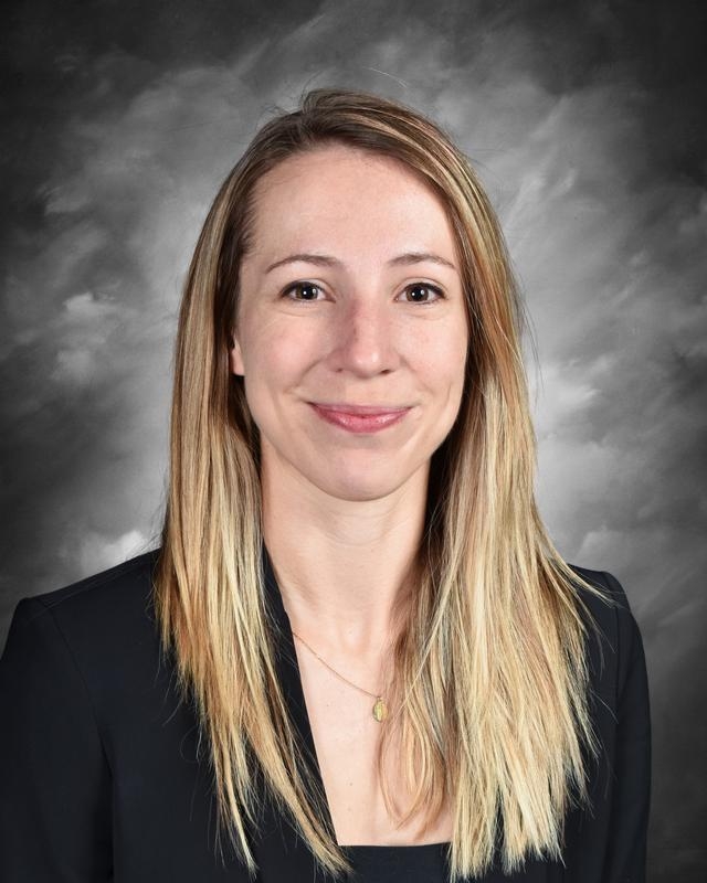 A smiling woman with long, straight hair wearing a black blazer, set against a cloudy background.
