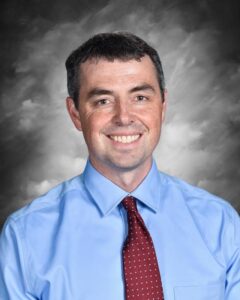 A smiling man with short hair, wearing a light blue shirt and a red tie, against a gray background.