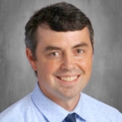A smiling man with short dark hair, wearing a light blue shirt and navy tie, against a gray background.