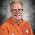Smiling man with glasses and short blonde hair, wearing an orange jacket with a logo, set against a gray background.