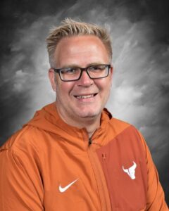 Smiling man with glasses and short blonde hair, wearing an orange jacket with a logo, set against a gray background.