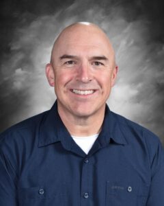 A smiling bald man in a navy shirt poses against a gray background. He looks friendly and approachable.