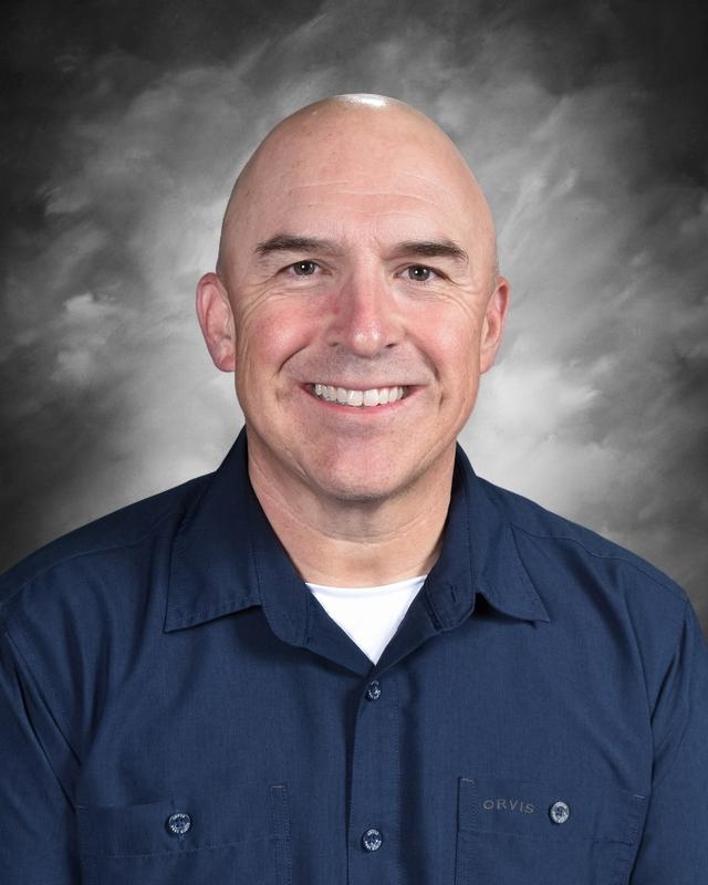 A smiling bald man in a navy shirt poses against a gray background. He looks friendly and approachable.