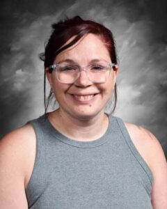 Smiling person with glasses and reddish-brown hair, wearing a gray sleeveless top, against a gray background.