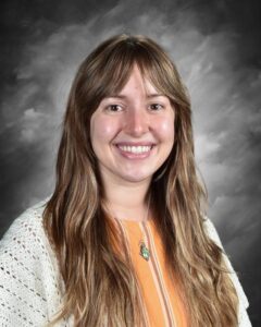 Smiling young woman with long, wavy hair, wearing an orange top and a white cardigan, set against a gray background.
