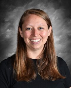 Smiling woman with long brown hair wearing a black shirt, against a gray backdrop.