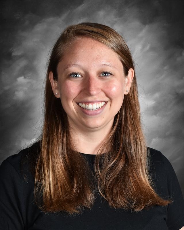 Smiling woman with long brown hair wearing a black shirt, against a gray backdrop.