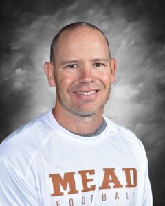 Portrait of a smiling man with short hair wearing a white shirt with "MEAD FOOTBALL" printed in orange.