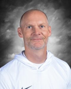 Portrait of a man with a shaved head and beard, wearing a white hoodie, smiling against a cloudy gray background.