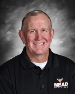 Smiling man wearing a black polo shirt with "MEAD WRESTLING" logo against a gray backdrop.