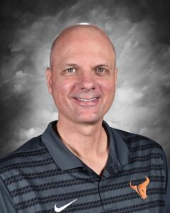 Smiling man with short hair wearing a dark striped polo shirt with an orange bull logo against a gray background.