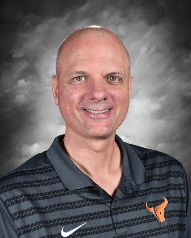 Smiling man with short hair wearing a dark striped polo shirt with an orange bull logo against a gray background.