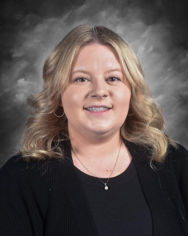 A smiling woman with wavy blonde hair, wearing a black top and cardigan, against a gray backdrop.