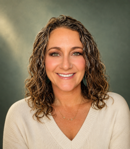 Smiling woman with curly hair, wearing a beige sweater and green earrings, set against a muted background.