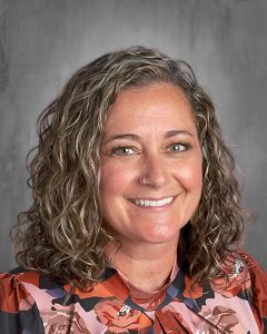 Smiling person with curly hair and a floral blouse against a gray background.