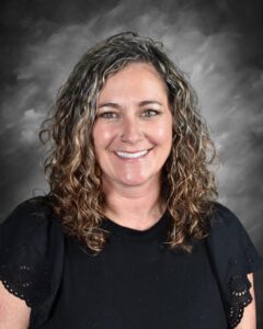 A smiling woman with curly hair wearing a black shirt, posed against a dark, textured background.