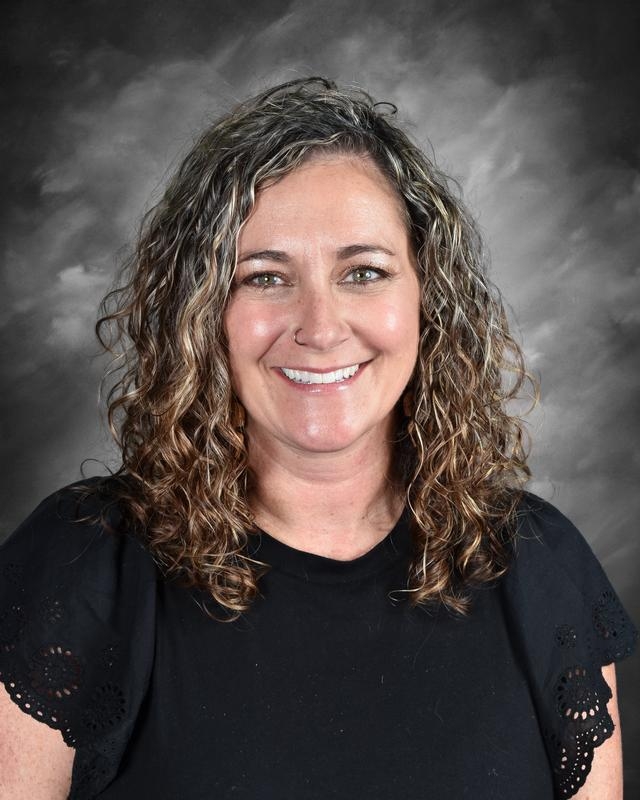 A smiling woman with curly hair wearing a black shirt, posed against a dark, textured background.