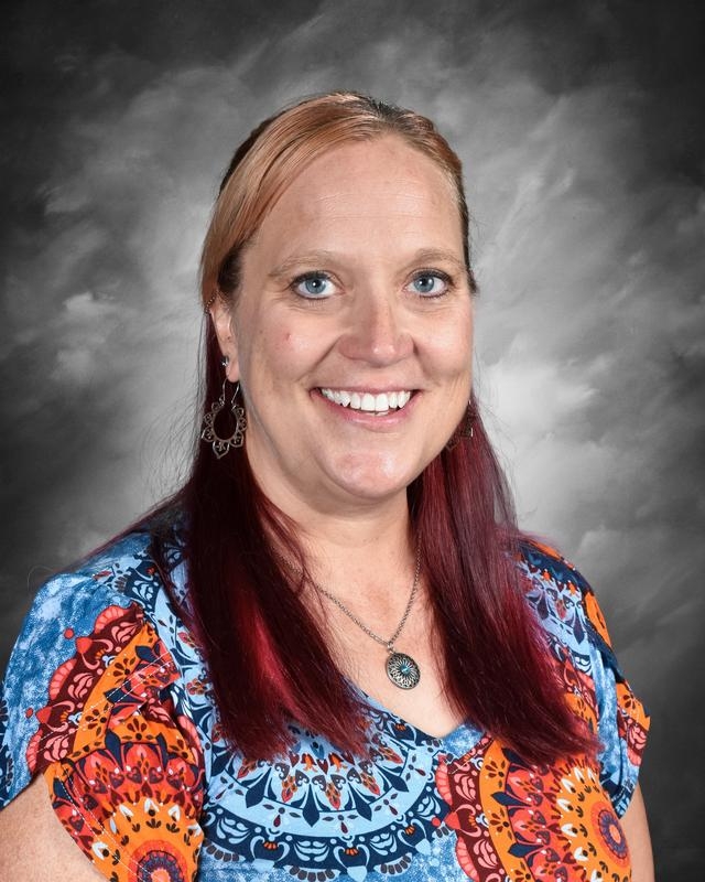 Smiling woman with long red hair, wearing colorful patterned clothing, against a gray background.