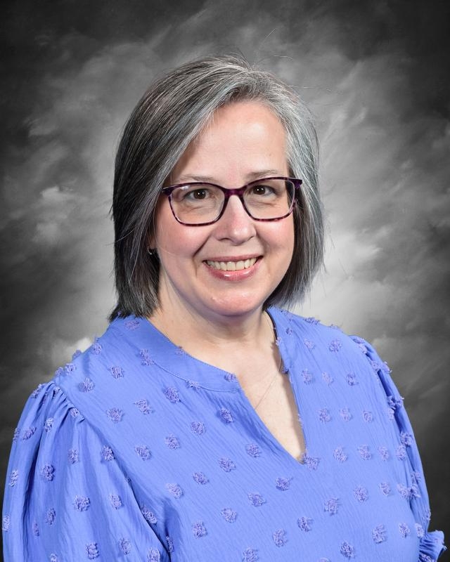 Smiling woman with gray hair and glasses, wearing a blue blouse, against a softly blurred gray background.