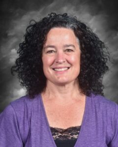 Smiling woman with curly black hair wearing a purple top and black lace underneath, set against a gray background.