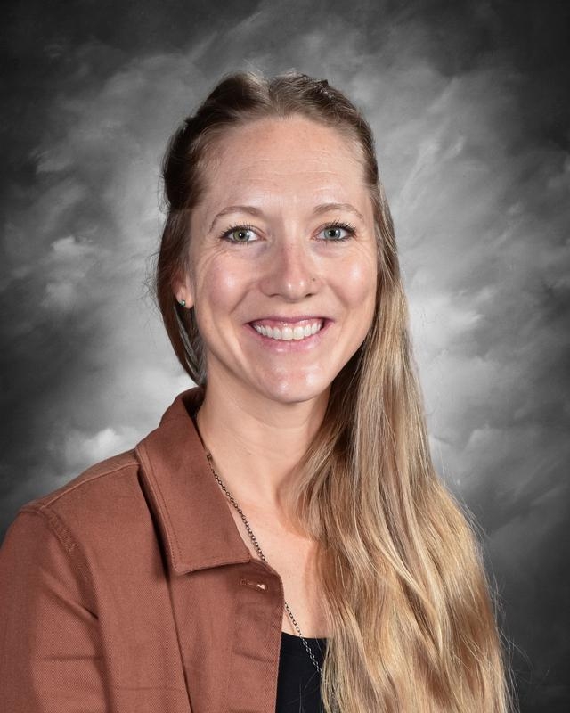 A smiling woman with long hair, wearing a brown jacket, poses against a gray background.