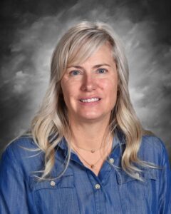 A woman with long, blonde hair smiles, wearing a denim shirt, against a soft gray backdrop.