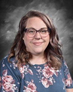 A smiling person with wavy hair wearing glasses and a floral shirt, set against a gray background.