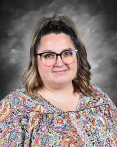 A smiling woman with shoulder-length hair, wearing glasses and a colorful patterned blouse, poses against a gray background.