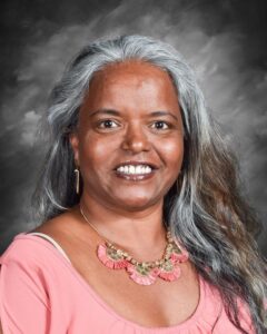 A smiling woman with long gray hair, wearing a pink top and a decorative necklace, against a dark background.