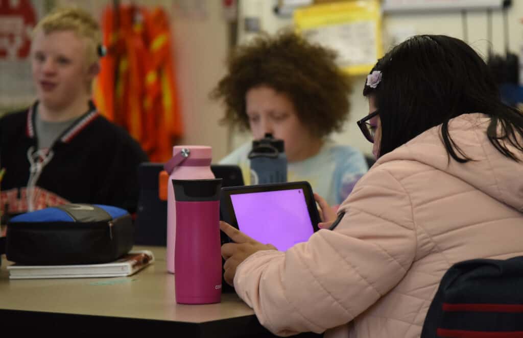A female high school student is sitting at a table working on an iPad with another male and female student at the same table in the background. 
