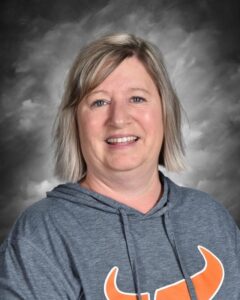 Una mujer sonriente con el pelo rubio corto, vestida con una sudadera gris con capucha y un dibujo naranja, sobre un fondo oscuro.