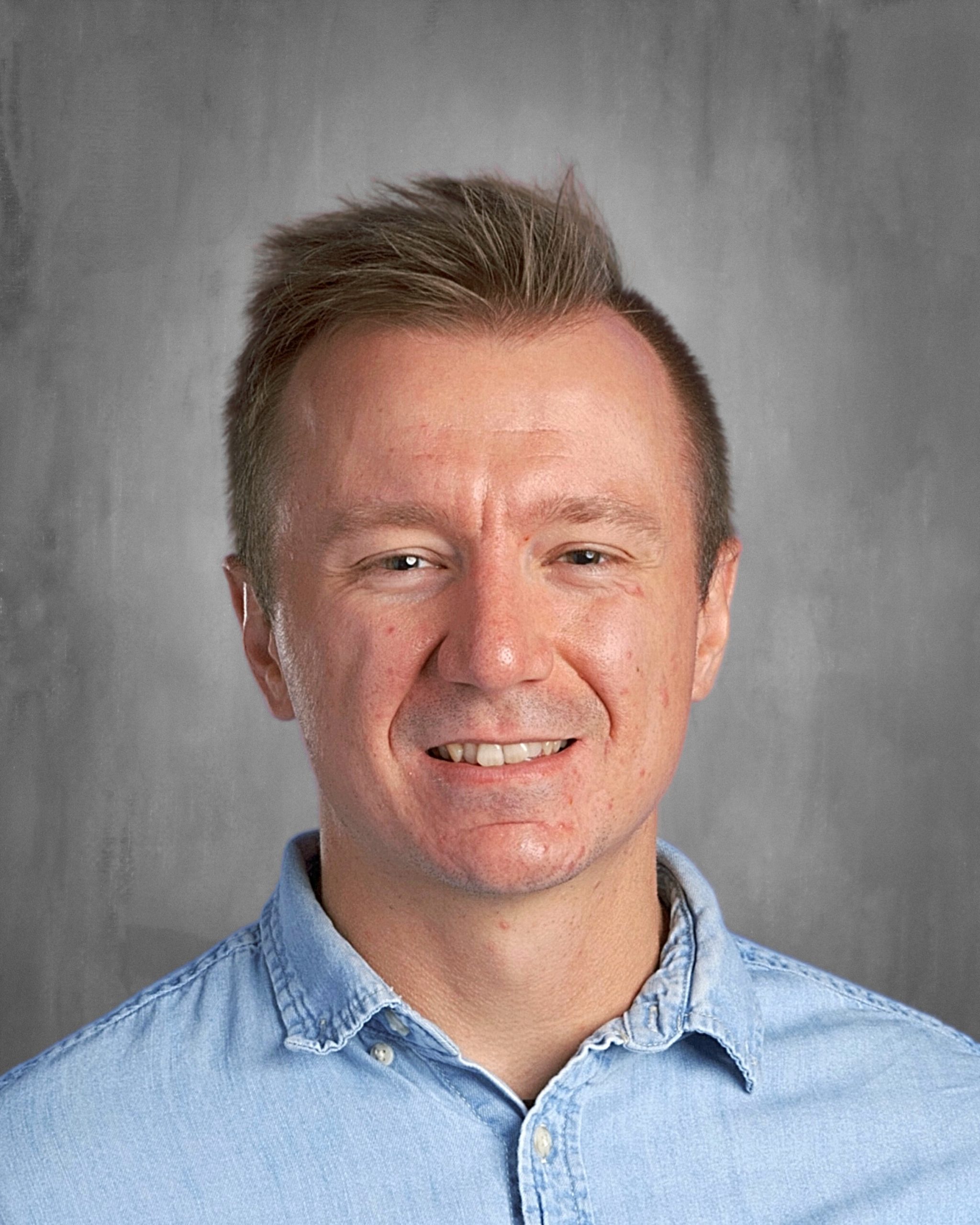 A person with short hair smiles, wearing a light blue denim shirt against a gray background.