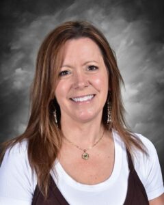 Smiling woman with long brown hair, wearing a white shirt and brown vest, against a gray background.