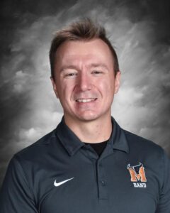 Smiling man in a dark polo with a band logo, against a gray background.