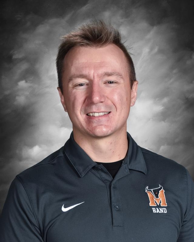 Smiling man in a dark polo with a band logo, against a gray background.