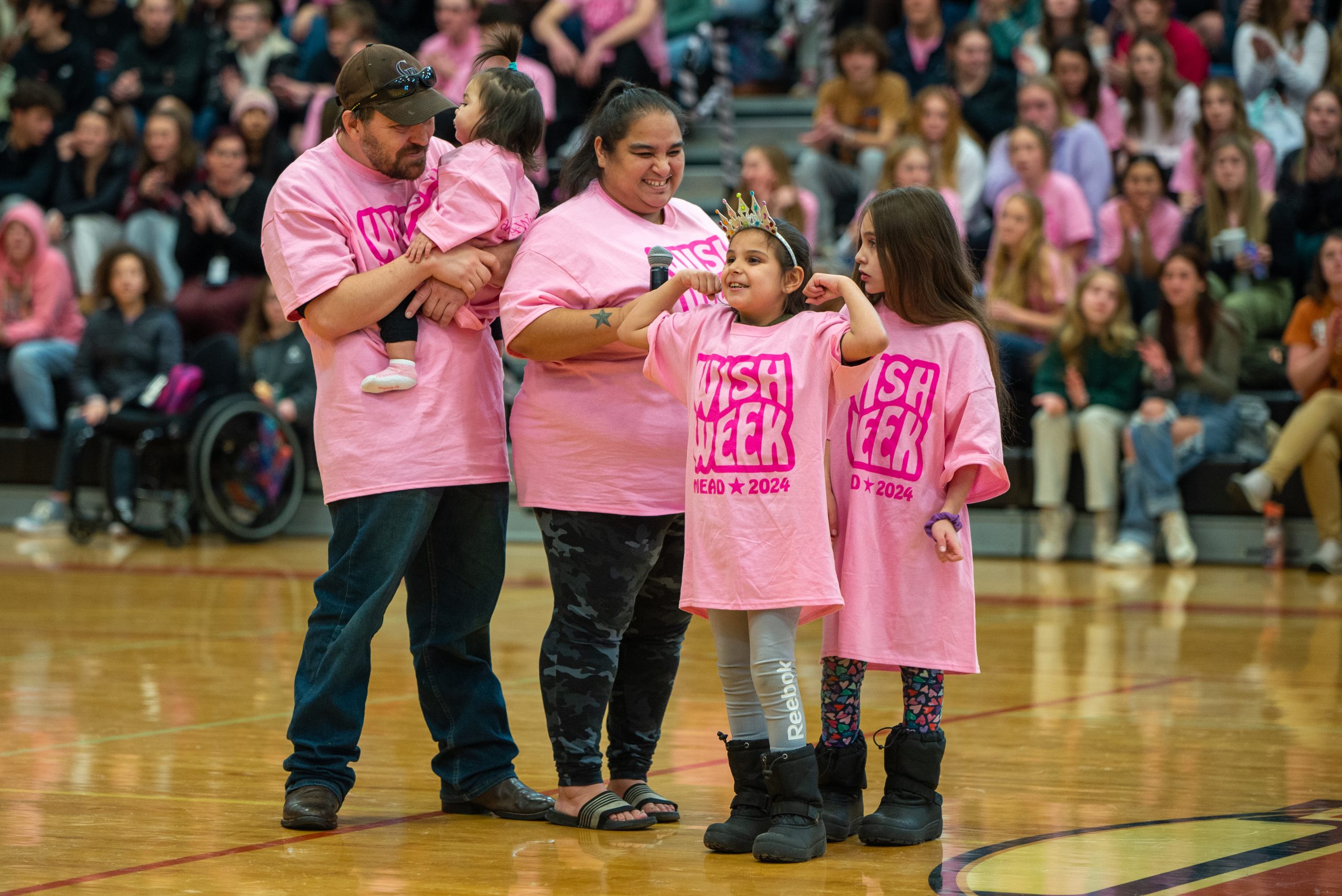 Familia de Make-a-Wish de pie en el gimnasio en Mead High con una multitud de estudiantes en las gradas borrosas detrás de ellos. El padre está a la izquierda con un niño en brazos, la madre a su lado a la derecha y las hijas a su lado. Llevan camisetas rosas a juego de la Semana de los Deseos 2024.