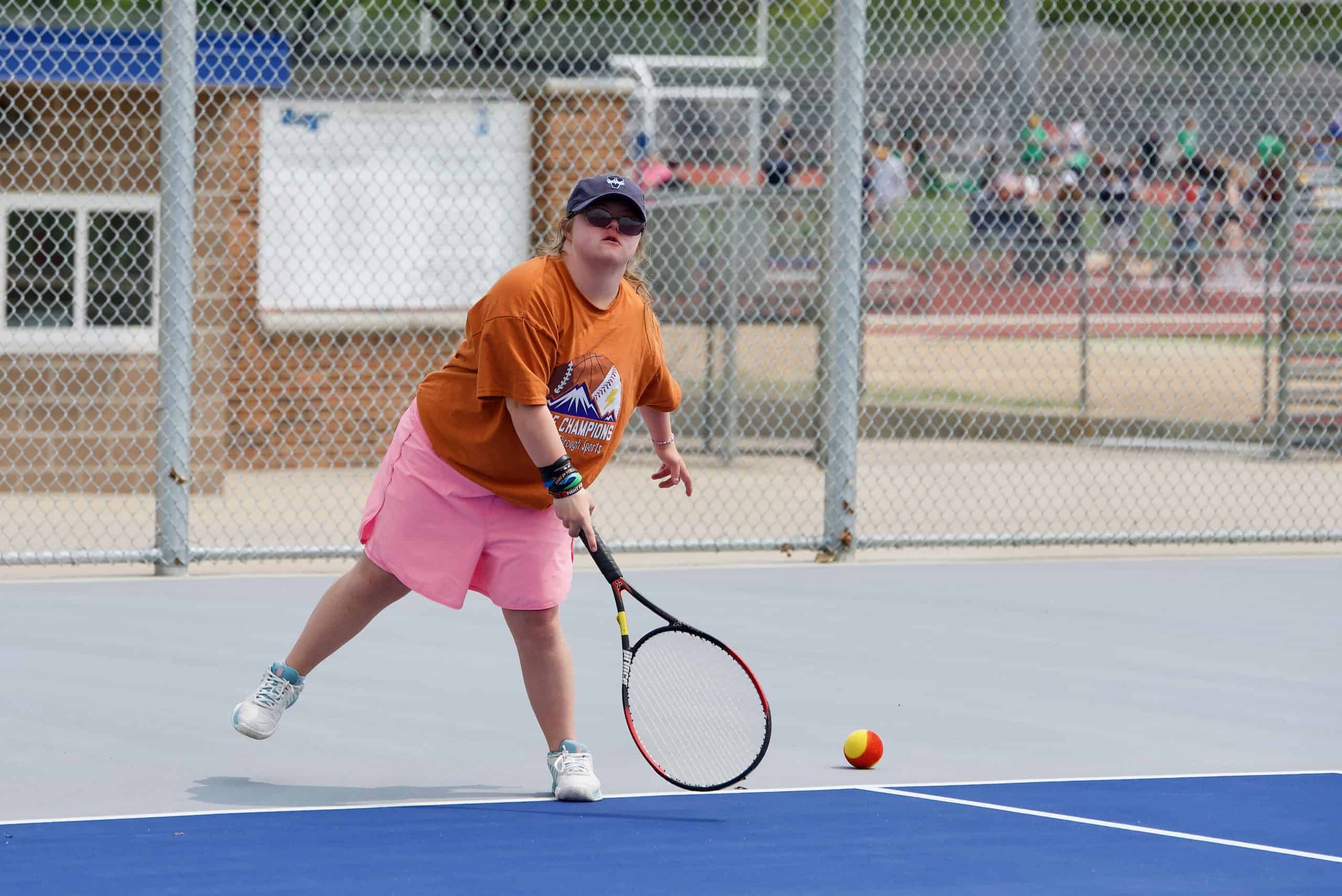 Female high school on a tennis court serving a tennis ball. She's wearing shorts, a Unified Day of Champions t-shirt, and a baseball hat.
