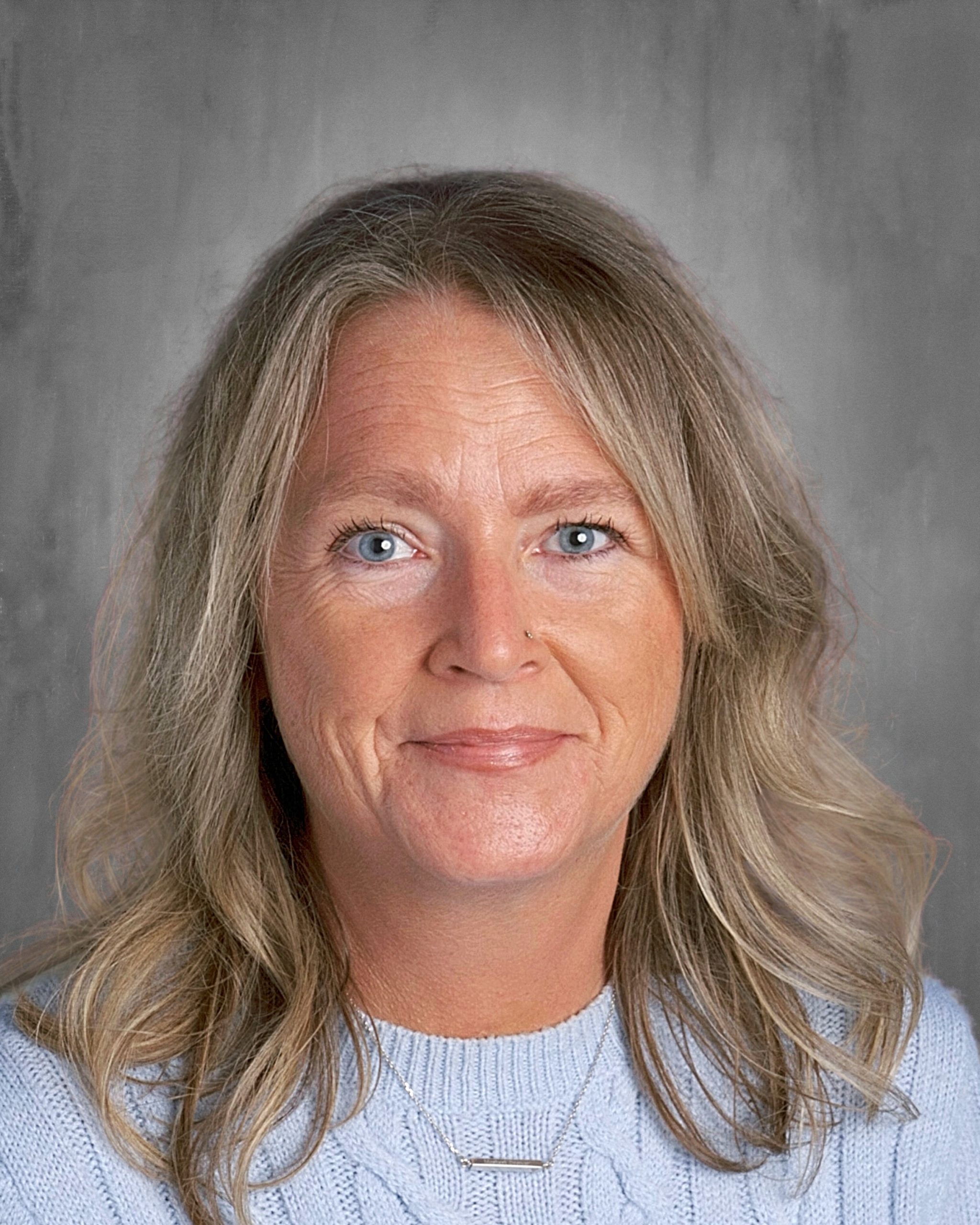 Woman with long blonde hair wearing a light blue sweater, smiling against a gray background.