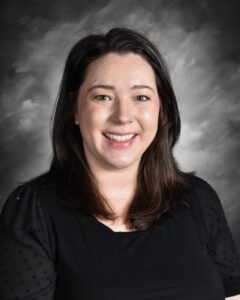 Smiling woman with long dark hair, wearing a black top, against a gray background.