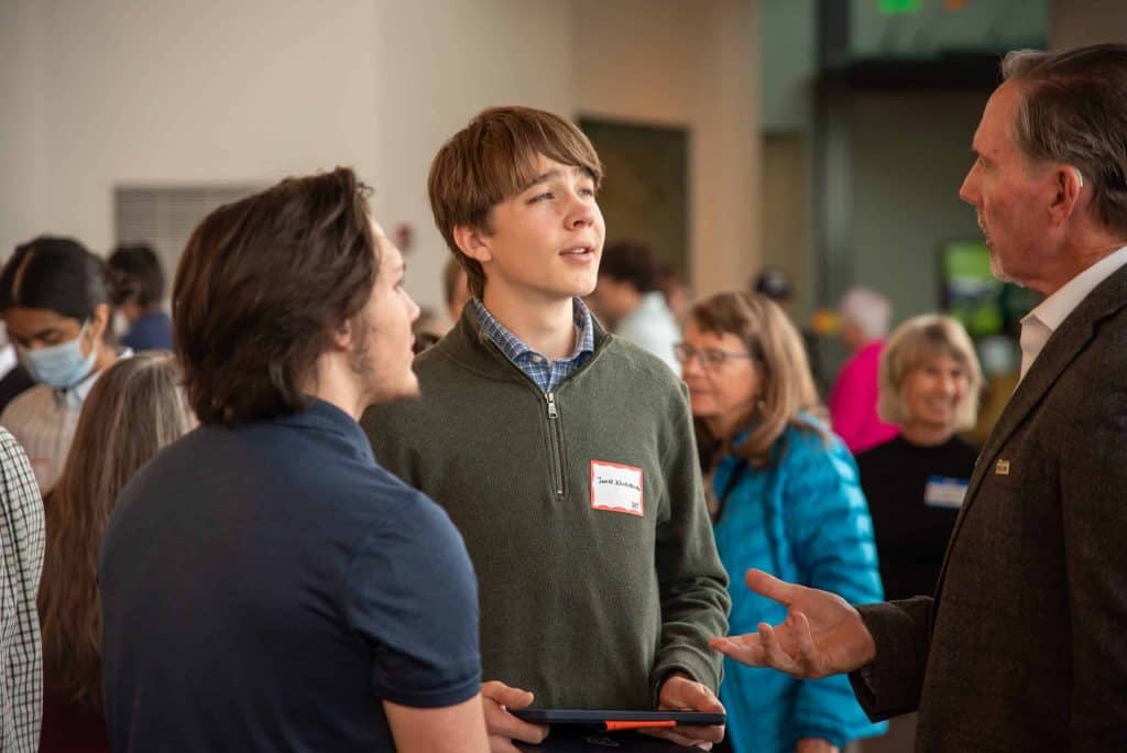 Two teenage boys are talking in a busy indoor event space with people gathered in the background.