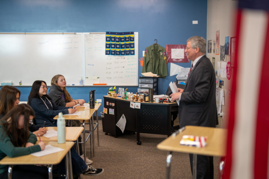 A Colorado supreme court justice stands at the front of a classroom while students listen attentively at their desks, taking notes.