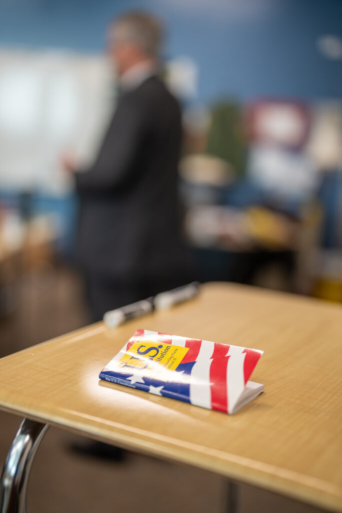 A book titled "U.S. Constitution" with a cover featuring the American flag lies on a desk in a classroom setting.