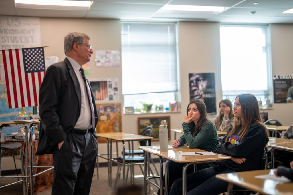 A Colorado supreme court justice stands in front of students in a classroom decorated with an American flag and various educational posters.