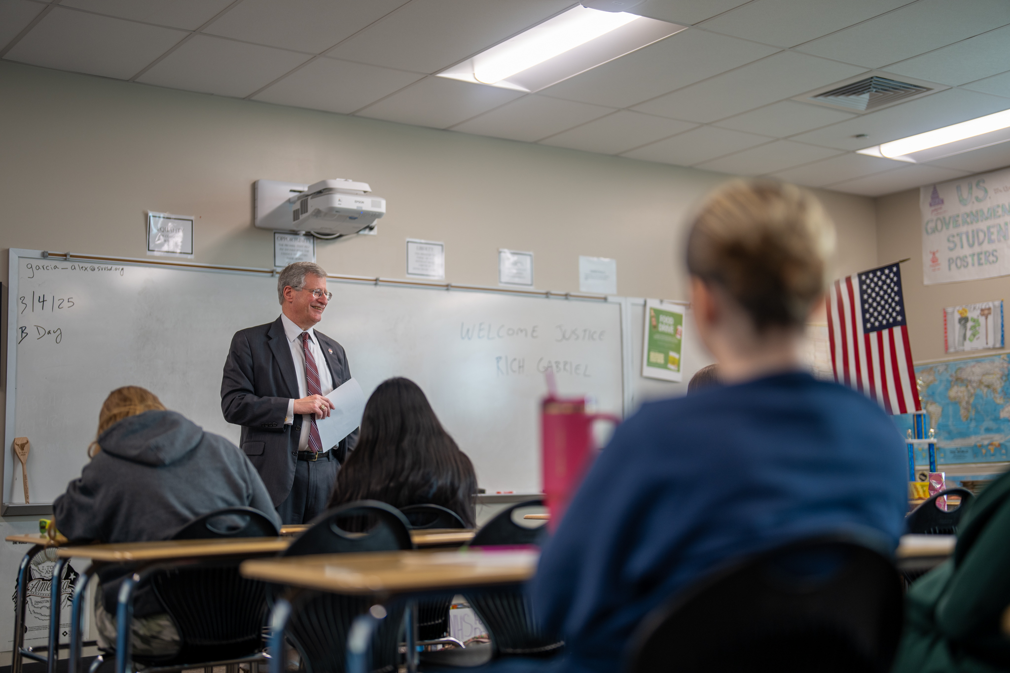 A judge speaks to students in a classroom, with a welcome sign behind him and students seated at desks.