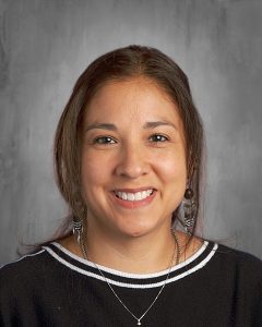 Smiling person with long hair, wearing a black top with white trim and earrings, against a gray background.