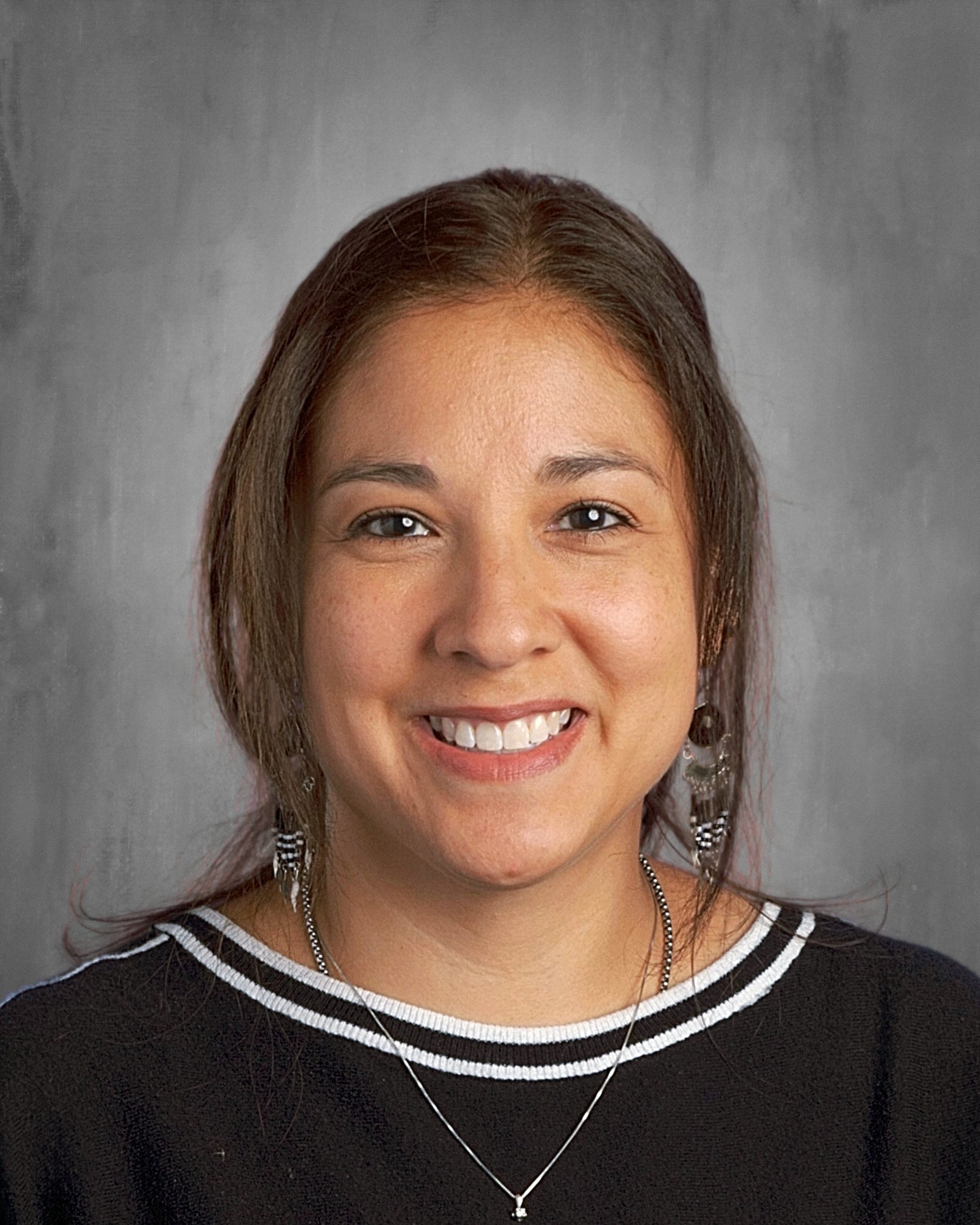 Persona sonriente con el pelo largo, que lleva una camiseta negra con ribete blanco y pendientes, sobre un fondo gris.