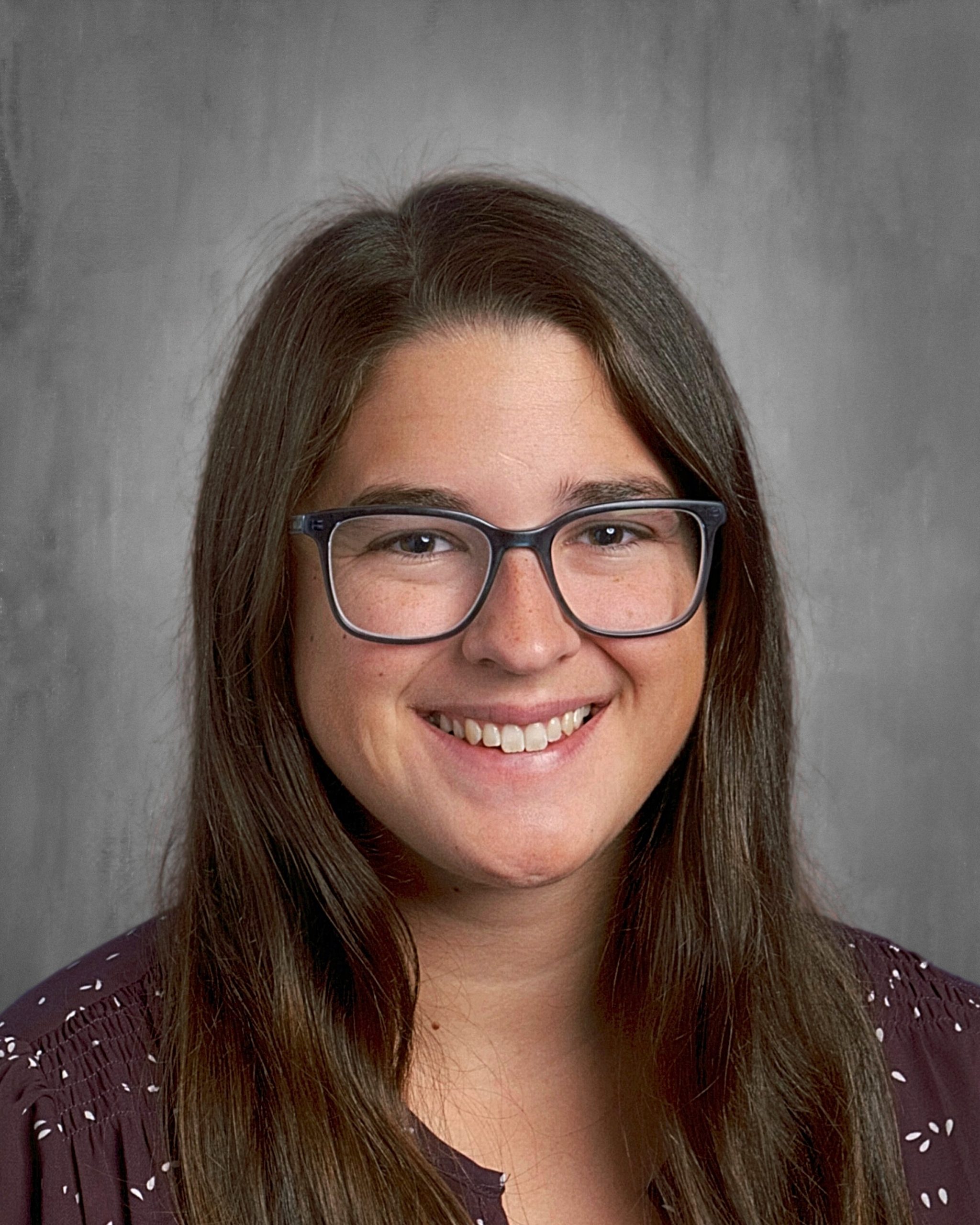 Smiling person with long brown hair and glasses, wearing a dark patterned top, against a gray background.